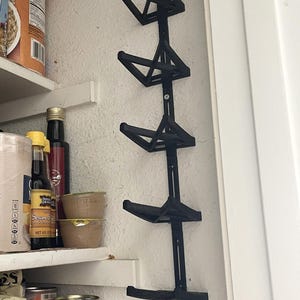 May include: Black, geometric, wall-mounted storage rack with multiple shelves. The rack is attached to a white wall and designed to hold items. The shelves are angled and appear to be made of plastic or metal.