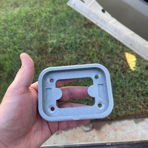 May include: A rectangular, silver-colored, 3D-printed mounting bracket with rounded corners. The component features a central rectangular cutout, two smaller rectangular indentations, and four circular screw holes positioned at each corner.