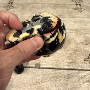 Peut inclure: Un objet en c&eacute;ramique arrondi pr&eacute;sentant un motif de gla&ccedil;ure marbr&eacute; avec des tourbillons de noir, de cr&egrave;me, de blanc et des touches d'orange. La surface a une finition brillante et r&eacute;fl&eacute;chissante avec des coulures de peinture irr&eacute;guli&egrave;res.