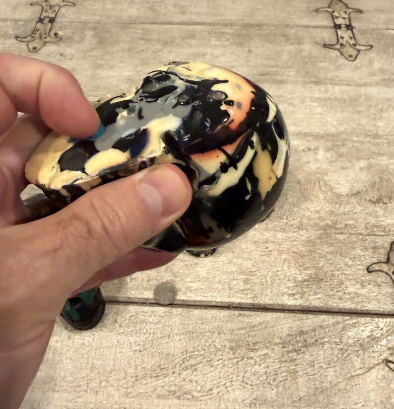 Peut inclure: Un objet en c&eacute;ramique arrondi pr&eacute;sentant un motif de gla&ccedil;ure marbr&eacute; avec des tourbillons de noir, de cr&egrave;me, de blanc et des touches d'orange. La surface a une finition brillante et r&eacute;fl&eacute;chissante avec des coulures de peinture irr&eacute;guli&egrave;res.