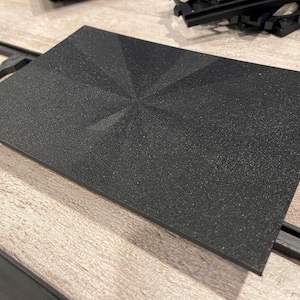 May include: A rectangular black panel with a textured surface and a geometric starburst pattern radiating from the center. The object features sharp, angular lines that create a faceted appearance across the flat, dark, matte-finished surface.