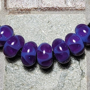May include: A necklace featuring seven oval-shaped glass beads. The beads are a deep purple with swirling patterns of lighter purple and blue. The beads are strung on a black cord, creating a striking contrast.