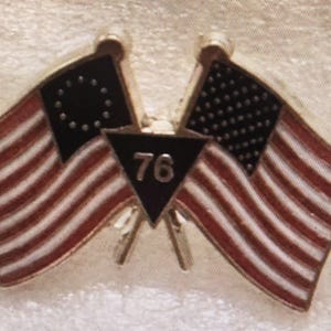 May include: A patriotic pin featuring two American flags with red and white stripes and a black field with white stars. A black triangle in the center displays the number "76". The pin has a silver-colored border and flagpoles.