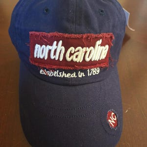 May include: Navy blue baseball cap with "north carolina" embroidered on a burgundy patch. The text "established in 1789" is below the patch. A small "NC" patch is on the side.