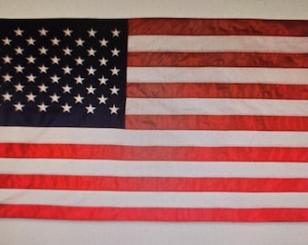 May include: A rectangular American flag with a blue canton filled with white stars. The flag has alternating red and white stripes. The flag is made of a slightly textured fabric and is hanging.