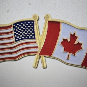 Op de afbeelding: Een goudkleurige speld met de gekruiste vlaggen van de Verenigde Staten en Canada. De Amerikaanse vlag heeft rode en witte strepen met een blauw veld met witte sterren. De Canadese vlag heeft een rood esdoornblad.