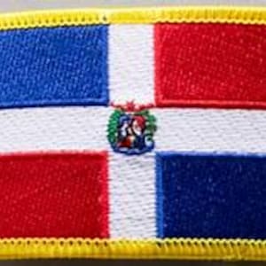 May include: Embroidered patch featuring the flag of the Dominican Republic. The rectangular patch has a white cross dividing the flag into four sections: blue, red, white, and red. A coat of arms is centered on the white cross. The patch is bordered with yellow stitching.