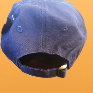 May include: A navy blue baseball cap with a curved peak. The cap has a brass-coloured adjustable buckle closure at the back. The cap is made of a durable fabric and features a classic six-panel design.