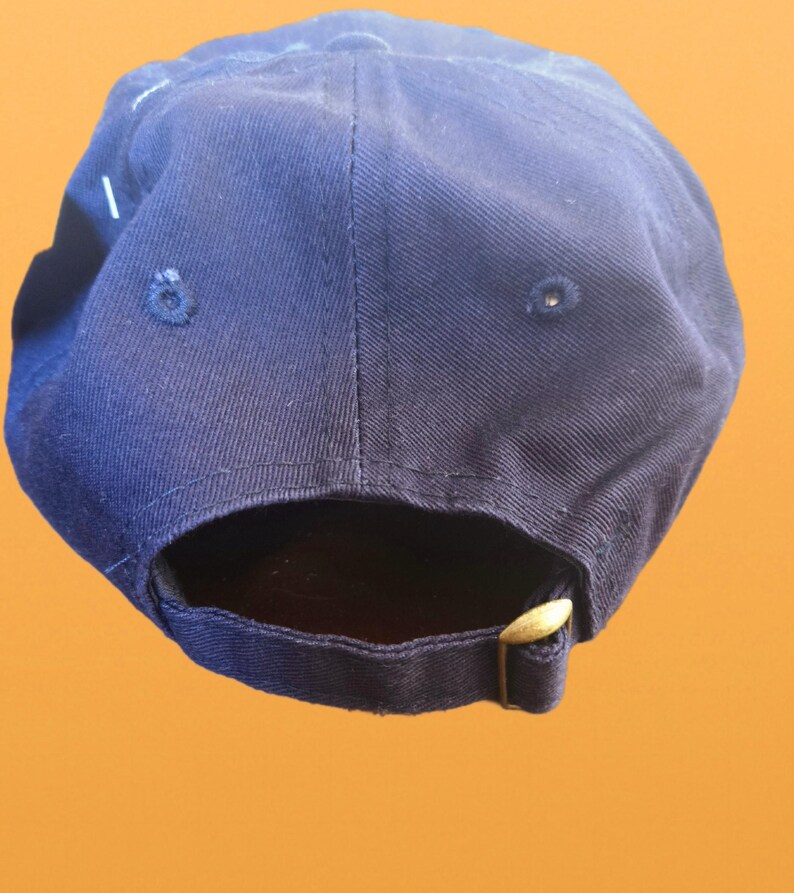 May include: A navy blue baseball cap with a curved peak. The cap has a brass-coloured adjustable buckle closure at the back. The cap is made of a durable fabric and features a classic six-panel design.