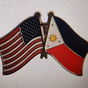 Op de afbeelding: Een speld met de vlaggen van de Verenigde Staten en de Filipijnen. De Amerikaanse vlag heeft rode en witte strepen met een blauw veld met witte sterren. De Filipijnse vlag heeft een witte driehoek, een gele zon en rode en blauwe secties.