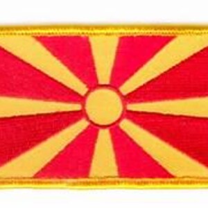 May include: Embroidered patch featuring the flag of North Macedonia. The rectangular patch has a red background with a yellow sunburst design emanating from a central circle. A yellow border frames the flag.