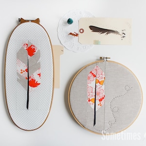 May include: Two embroidery hoops with fabric stretched across them. The fabric is a light gray color and features a red and white floral pattern. The floral pattern is in the shape of a feather. The embroidery hoops are hanging on a white wall. The text "Sometimes Crafter" is visible in the bottom right corner of the image.