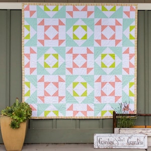 May include: A quilt with a patchwork design featuring pink, green, and white squares. The quilt is hanging on a wall with a wooden sign that reads "Garden Sweet Garden".