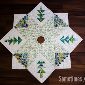 May include: A white, green and yellow quilted Christmas tree skirt with a snowflake design and a hole in the centre for a tree trunk. The skirt is made of fabric with a white background and a pattern of small green and yellow leaves. The skirt is surrounded by a white border.
