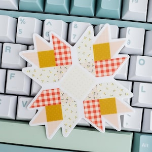 May include: A star-shaped sticker with a patchwork quilt design. The sticker features a mix of patterns, including gingham, floral, and solid squares in shades of peach, yellow, and red. It rests on a light blue and white keyboard.