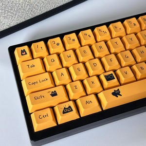 May include: A yellow mechanical keyboard with black keycaps featuring various cat-themed illustrations. The keyboard has a black frame and a black and white cable.