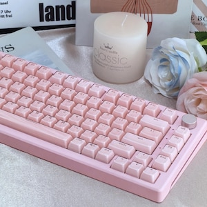 Op de afbeelding: Een roze draadloos toetsenbord met witte toetsen en een ronde zilveren knop rechtsboven. Het toetsenbord ligt op een wit oppervlak met een witte kaars en roze bloemen.