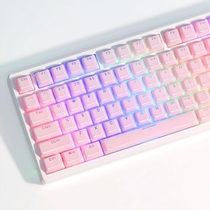 Op de afbeelding: Een roze en wit toetsenbord met regenboogkleurige LED-achtergrondverlichting. De toetsen hebben witte letters en zijn gerangschikt in een standaard QWERTY-indeling.
