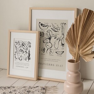 Op de afbeelding: Twee ingelijste kunstdrukken met botanische illustraties en tekst, &eacute;&eacute;n met Genesis 1:29 en de andere met Spreuken 15:17. Een roze vaas bevat een gedroogd palmblad. De achtergrond is een lichtbeige muur en een getextureerd tapijt.