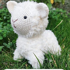 May include: A white crocheted lamb sitting in green grass. The lamb has a fluffy texture and black eyes. The text "Crochet pattern" is visible at the bottom of the image.