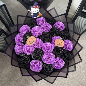 Può includere: Un bouquet di rose artificiali in tonalità alternate di viola e nero, avvolto in carta scura traslucida. La composizione presenta ritagli decorativi di un personaggio Hello Kitty in costume da strega e due zucche di colore arancione.