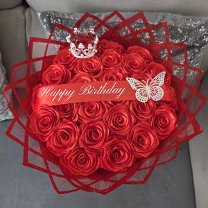 Peut inclure: Un bouquet de roses rouges, disposées en cercle, avec un ruban rouge portant l'inscription "Happy Birthday". Une petite couronne argentée et un papillon blanc ornent le dessus. Le bouquet est enveloppé dans un matériau rouge à motifs géométriques.