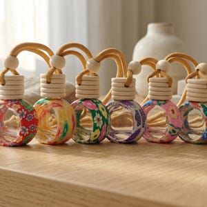 May include: Six small, round, clear glass car air freshener bottles arranged in a row. Each bottle features a unique colourful floral pattern on the glass and is topped with a light-coloured wooden cap and a tan hanging cord with a wooden bead.
