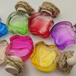 May include: A collection of seven small, apple-shaped glass perfume bottles in blue, green, red, purple, clear, pink, and yellow. Each bottle features a wooden cap wrapped in thick, tan rope with subtle gold metallic threading throughout the cord.