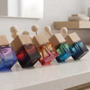 May include: Five square glass car air freshener bottles with wooden caps and spherical wooden toppers. Each bottle features a distinct gradient colour including purple, red, orange, green, and blue, arranged in a row on a flat surface.