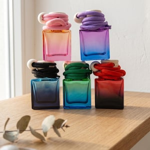May include: Five square glass bottles with gradient colours, each topped with a wooden cap and a thick, coiled rope in a matching hue. The bottles feature vibrant colour transitions, including pink, purple, black, blue, and red, arranged in a stack.