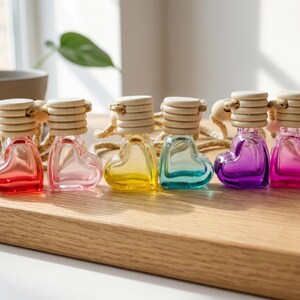 May include: A set of six heart-shaped glass bottles with light-coloured wooden caps and attached hanging cords. The bottles are filled with liquids in various colours including red, light pink, yellow, light blue, purple, and bright pink.