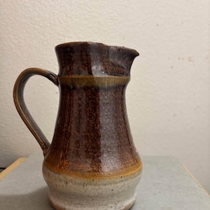 Puede incluir: Jarra de cerámica hecha a mano con un esmalte marrón oscuro y una base crema. La jarra tiene un asa curva y un pico para verter. La cerámica es probablemente gres, con un diseño rústico y funcional.