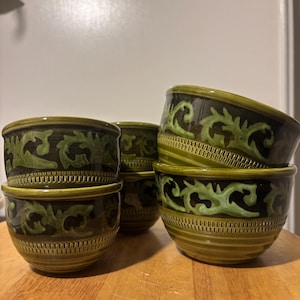 May include: A stack of six green ceramic bowls with a dark brown and green floral design. The bowls have a ribbed texture on the lower half and a decorative band around the top. The bowls are stacked on a wooden surface.