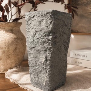 May include: A tall, rectangular vase with a rough, dark gray, stone-like texture. The vase has a wide, flat top opening and tapers slightly toward the base, featuring a rugged, uneven surface that mimics natural rock formations.