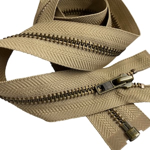 May include: A beige zipper with brass teeth and a brass slider. The zipper is folded in half and the slider is in the middle of the zipper. The zipper is made by YKK.