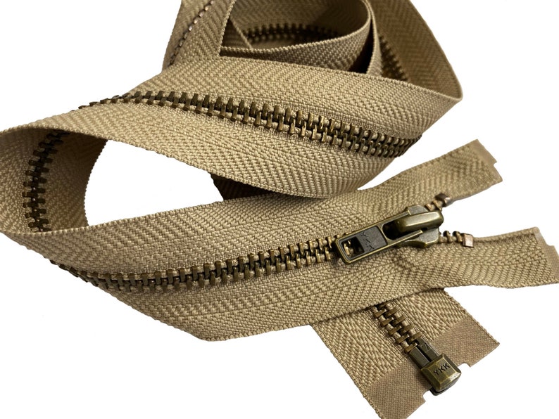May include: A beige zipper with brass teeth and a brass slider. The zipper is folded in half and the slider is in the middle of the zipper. The zipper is made by YKK.