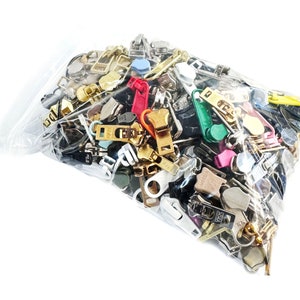 Puede incluir: Una bolsa de plástico transparente llena de tiradores de cremallera de varios colores y metálicos. Los tiradores son de diferentes formas y tamaños, incluyendo algunos con dientes y otros sin.