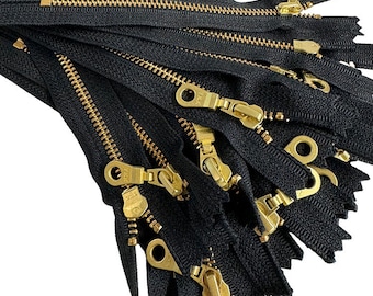 May include: A collection of black zippers with gold-colored metal teeth and pulls. The zippers are arranged in a slightly overlapping pattern, showcasing the details of the metal components and the fabric tape.