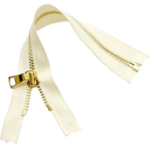 May include: A cream-colored zipper featuring metallic gold teeth and a matching gold pull tab. The fabric tape has a visible herringbone weave pattern, and the metal components have a polished, reflective finish against the light fabric.