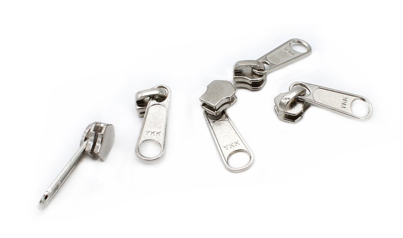 May include: Five silver zipper pulls with the YKK logo. The zipper pulls are isolated on a white background.