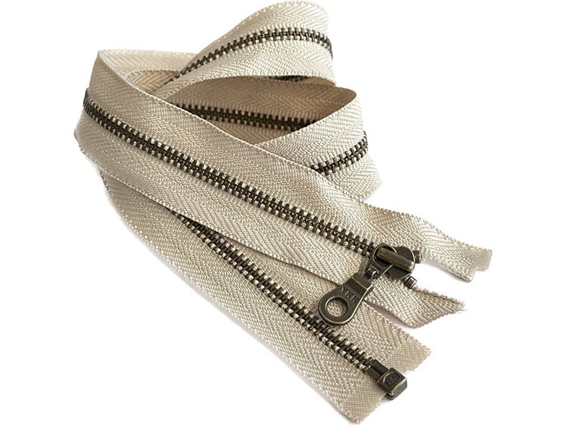 May include: A beige zipper with antique brass teeth and a slider. The zipper is folded in half and the slider is at the top of the fold.