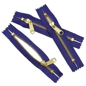 May include: Three dark purple zippers with metallic gold-toned teeth and matching gold-toned metal sliders. Each slider is clearly embossed with the YKK brand logo. The zippers are arranged in a scattered pattern against a plain white background.