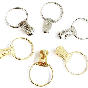 Puede incluir: Seis tiradores de cremallera con anillos metálicos. Los tiradores son plateados y dorados con el nombre de la marca YKK impreso en ellos.