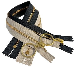Könnte beinhalten: Zwei Reißverschlussbänder, eines beige und eines schwarz, mit goldenen Metallzähnen und Schiebern. Die Reißverschlüsse sind ineinander verschlungen und die Schieber sind mit einem Metallring verbunden.