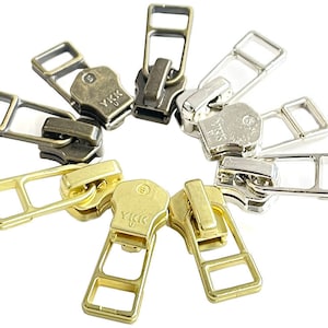 May include: A collection of zipper pulls in various metallic finishes, including gold, silver, and bronze. Each pull features a rectangular handle and a branded YKK zipper head. The pulls are arranged in a circular pattern against a white background.