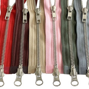 Puede incluir: Ocho cremalleras metálicas en varios colores, incluyendo rojo, marrón, beige, rosa, verde y gris. Las cremalleras tienen un acabado plateado y están todas cerradas. Las cremalleras están etiquetadas con la marca "YKK".