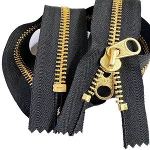 May include: A close-up of a black zipper with gold metal teeth and a gold slider. The zipper is folded in half and the slider is in the closed position.