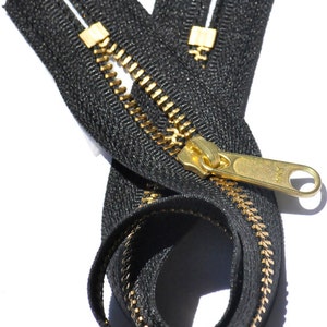 May include: Black zipper with gold-colored teeth and pull tab. The zipper is open, with the teeth visible. The zipper tape is black. The pull tab is a shiny gold color. The zipper is on a white background.