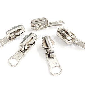May include: Five silver zipper pulls with a YKK logo. The zipper pulls are arranged in a circular pattern on a white background.
