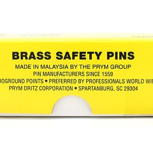 May include: A yellow box with black text that reads "Brass Safety Pins" and "Made in Malaysia by the Prym Group". The box also states "Pin Manufacturers Since 1559", "Microground Points", "Preferred by Professionals World Wide", "Prym Dritz Corporation", and "Spartanburg, SC 29304".